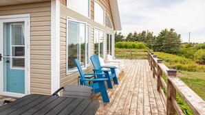 A wooden deck with blue Adirondack chairs and a table, overlooking a lush landscape. - 3-Bedroom Cavendish Oceanfront Cottage - Located inside National Park (North Rustico)