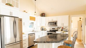 A modern kitchen with white cabinets, stainless steel appliances, a granite countertop, and wicker chairs. - 3-Bedroom Cavendish Oceanfront Cottage - Located inside National Park (North Rustico)
