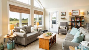 A spacious living room with large windows, a fireplace, a bookshelf, and a comfortable seating area. - 3-Bedroom Cavendish Oceanfront Cottage - Located inside National Park (North Rustico)