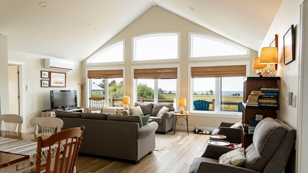 A spacious living room with a high ceiling, large windows, and a view of the outdoors. - 3-Bedroom Cavendish Oceanfront Cottage - Located inside National Park (North Rustico)