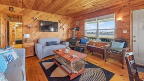 A cozy cabin interior with a wooden ceiling, a flat-screen TV mounted on the wall, a sofa, a chair, a wooden coffee table, and a large window offering a view of the outdoors.