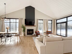 A spacious living room with a high ceiling, a fireplace, a large sofa, and a dining area with a table and chairs. - Modern Chalet w/ Hot Tub minutes to Cavendish - Seawood Haven (North Rustico)