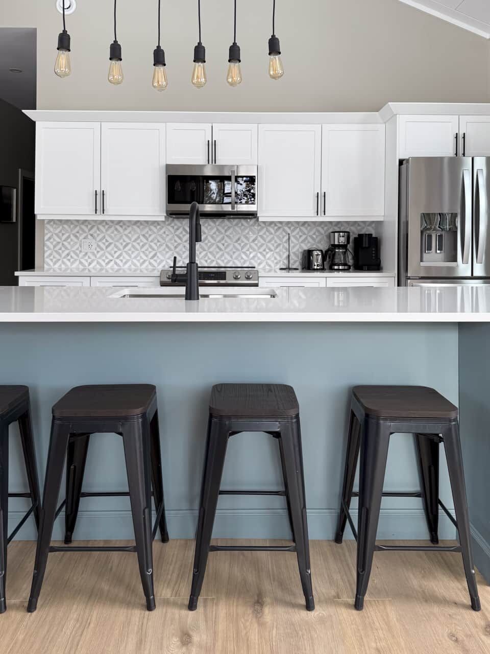 A modern kitchen with white cabinets, a stainless steel refrigerator, a built-in oven, and a bar area with stools.
