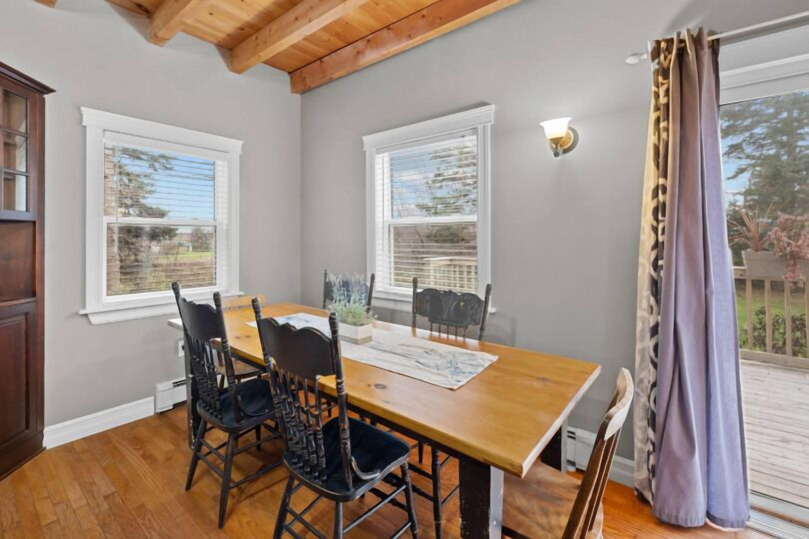 A dining area with a wooden table and chairs, two windows with blinds, a ceiling with exposed beams, and a view of the outdoors through a curtain.
