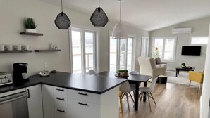 Een moderne keuken met witte kasten, een zwart aanrecht en een koffiezetapparaat. Er is een eethoek met een tafel en stoelen, en een woonkamer met een bank en een televisie.