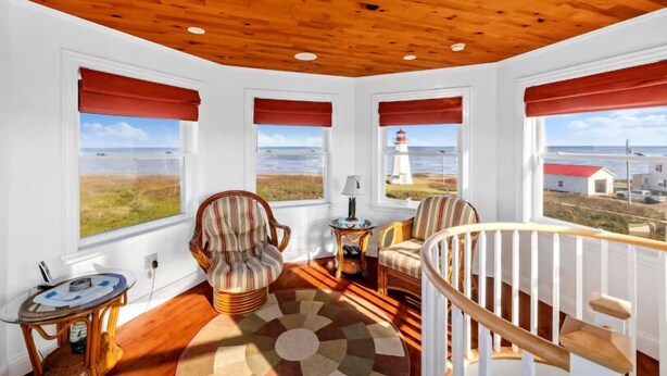 Une pièce chaleureuse avec un plafond en bois, de grandes fenêtres dotées de stores rouges, un tapis à motifs et une vue sur la mer et un phare.