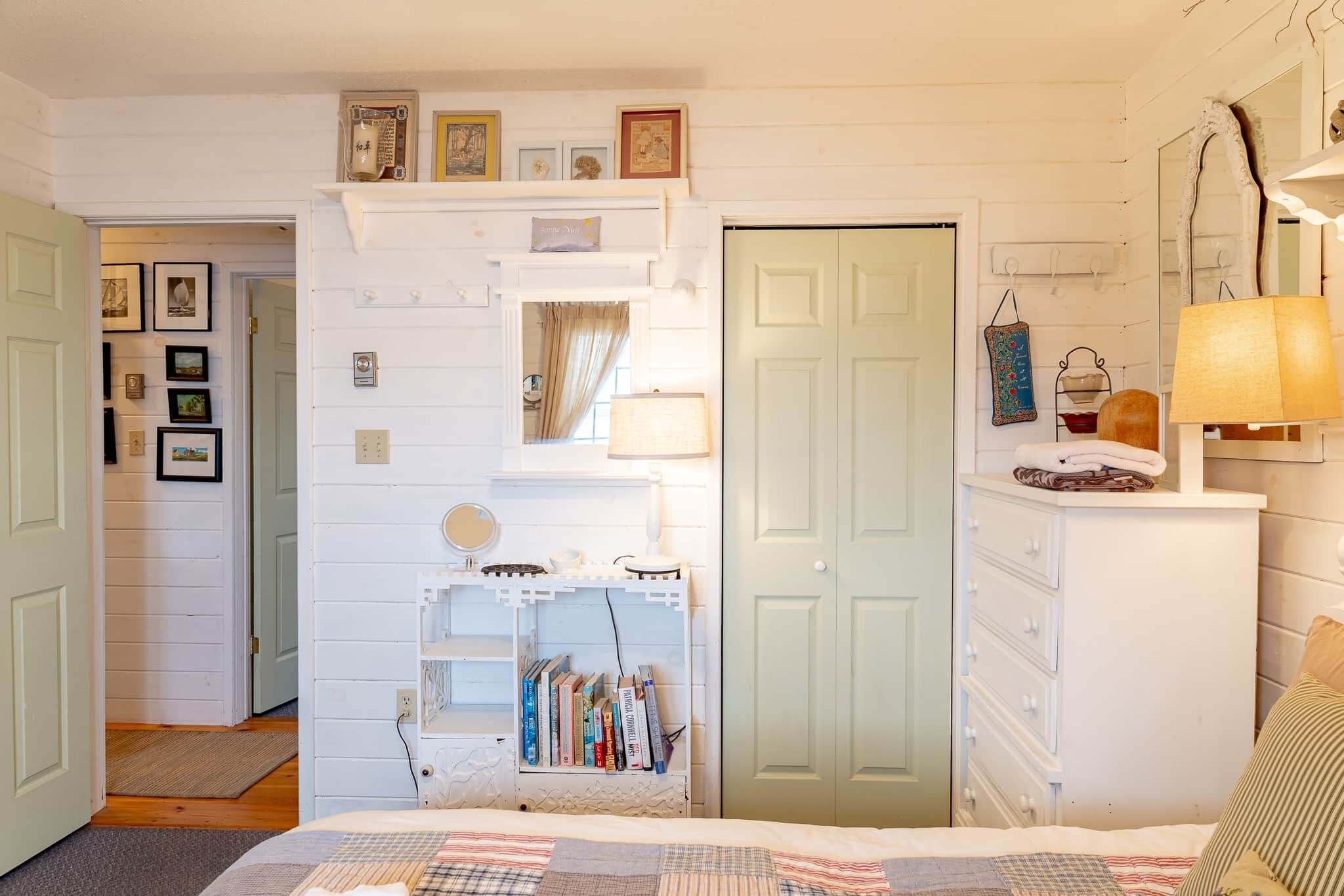 A bedroom with a bed, a dresser, a vanity with a mirror, a shelf with books, and a door.