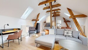 Un loft moderno con techo inclinado, vigas de madera expuestas, un sofá seccional gris, un escritorio con silla y una kitchenette al fondo.