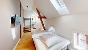 Un dormitorio con techo inclinado, tragaluces, cama, piso de madera y vista a un edificio de ladrillo.
