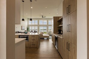 A modern kitchen with wooden cabinets, a central island, pendant lighting, and a view through large windows.