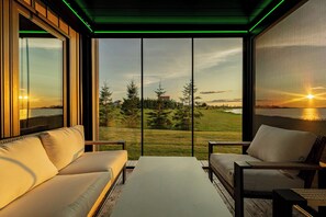 A room with a sofa and chairs facing a large window with a view of a sunset over a grassy landscape.