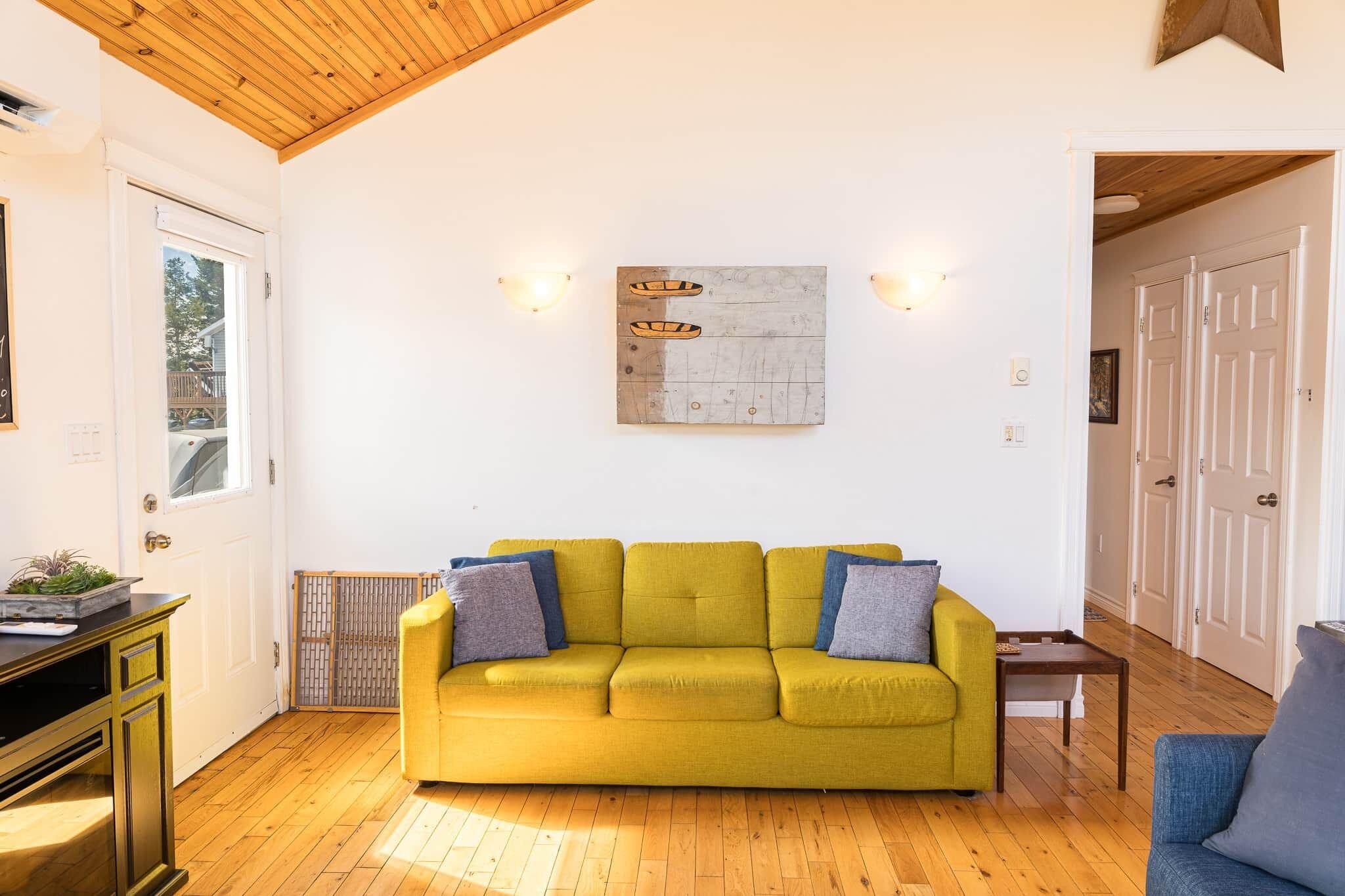 Un salon lumineux avec un canapé jaune, une petite table et une œuvre d’art accrochée au mur.