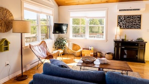 A cozy living room with a wooden ceiling, a large window, a flat-screen TV, and a wall-mounted chalkboard with writing. - Riverside Summer Retreat on PEI with Hot Tub, Deck, Kayaks & Pet-Friendly Stay (Prince Edward Island)