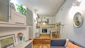 A compact kitchen with white cabinets, a stove, and a sink. There is a dining area with a table and chairs, and a grey sofa with a cushion.