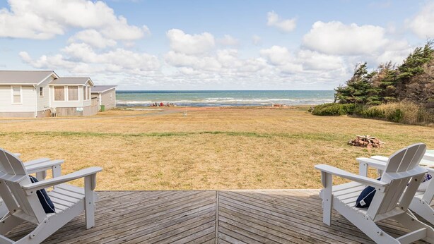 Une vue sur la plage depuis une terrasse en bois, avec deux chaises blanches, un petit bâtiment et un ciel dégagé.
