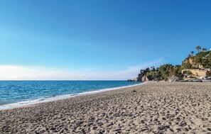 Ein weiter, sandiger Strand mit sanften Wellen, einem wolkenlosen blauen Himmel und einem felsigen Klippenhang mit Vegetation rechts.
