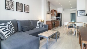 Ein modernes Wohnzimmer mit einem grauen Sofa, einem Marmortisch und einem Essbereich mit einem Holztisch und Stühlen. Im Hintergrund ist eine Küche mit Herd und Schränken zu sehen.