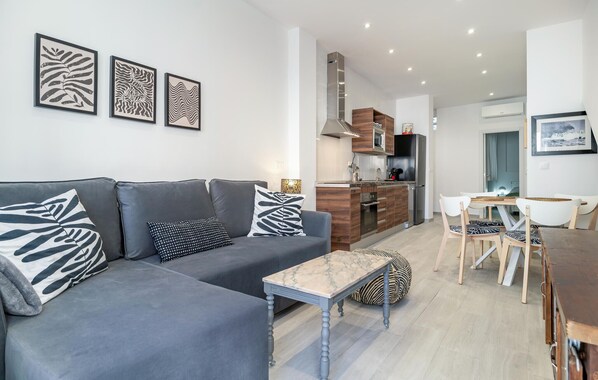 Un salon moderne avec un canapé gris, une table basse en marbre et un coin repas comprenant une table et des chaises en bois. On aperçoit une cuisine avec une cuisinière et des placards au fond.