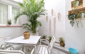 Un petit espace patio avec une table et des chaises blanches, une grande plante en pot et des éléments décoratifs suspendus au mur.