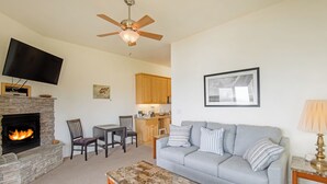 A living room with a stone fireplace, a grey sofa, a coffee table, a dining table with chairs, a ceiling fan, and a kitchen in the background. - Seagull by OceanviewHotTubs Oceanfront Tesla charging Oceanfront Shelter Cove (Whitethorn)