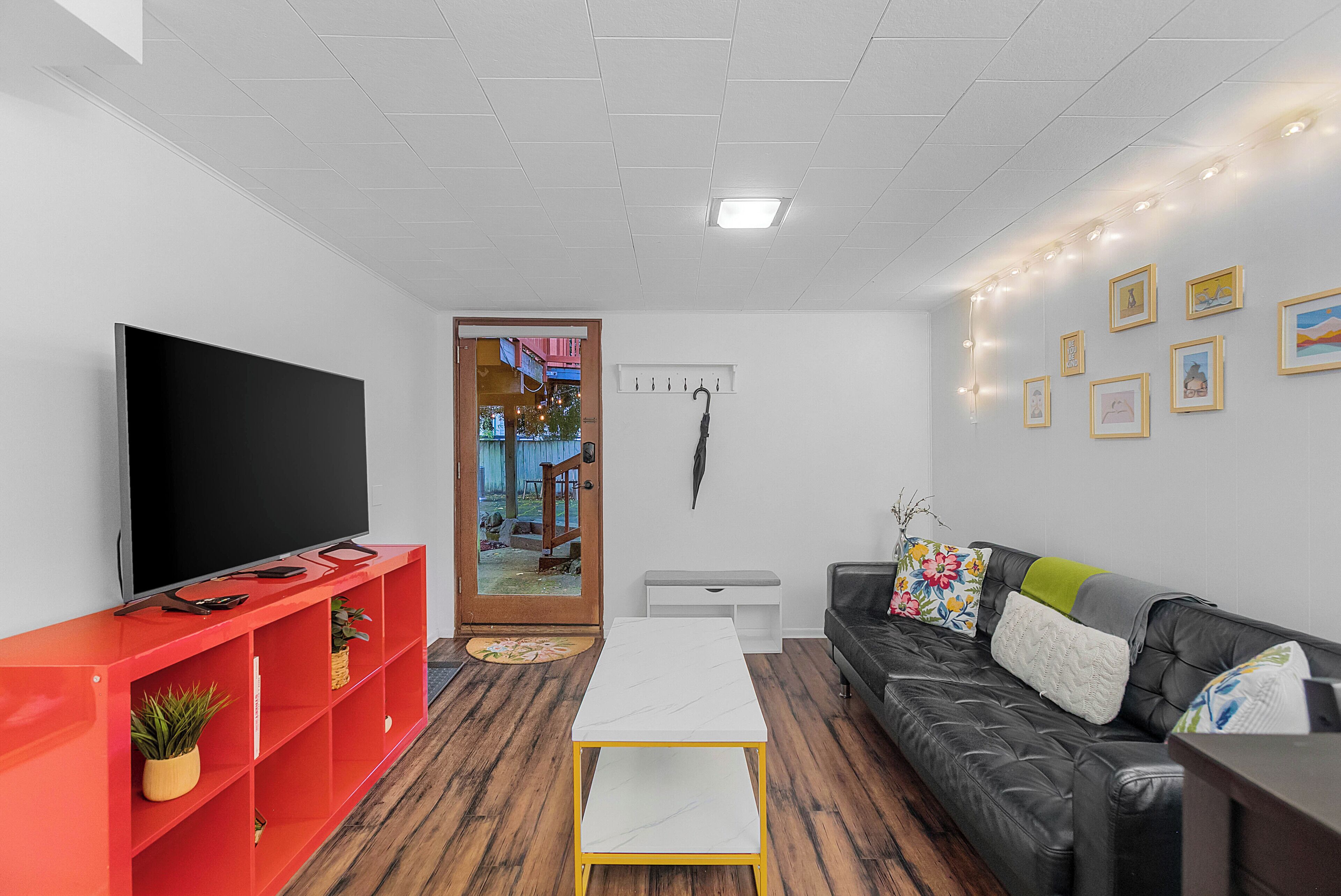 A modern living room with a flat-screen TV, red shelving unit, a white coffee table, a black leather sofa, and a door leading to an outdoor area.