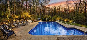 A swimming pool with lounge chairs and a deck, surrounded by trees and a sunset.