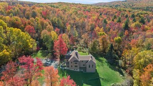 A large, single-story house with a symmetrical roof and a central chimney, surrounded by a well-maintained lawn and dense forest with autumn foliage.
