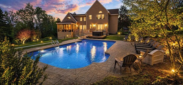A swimming pool with a deck, surrounded by outdoor furniture and a large house.