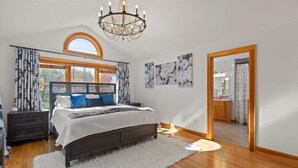 A bedroom with a large bed, a chandelier, a window with curtains, and a mirror reflecting a bathroom.