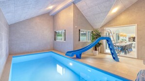 Une piscine intérieure moderne, dotée d’un toboggan bleu, de grandes fenêtres et d’un espace repas visible à travers des portes vitrées.