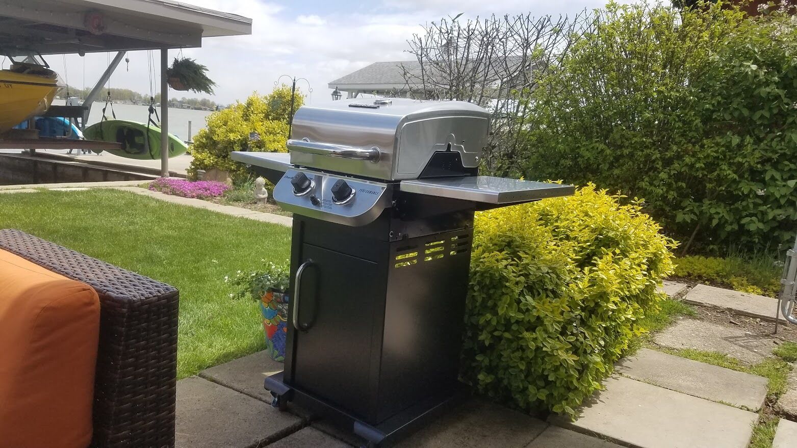 A portable gas grill with two burners, a side shelf, and a control panel, set up outdoors with a grassy lawn and bushes in the background.