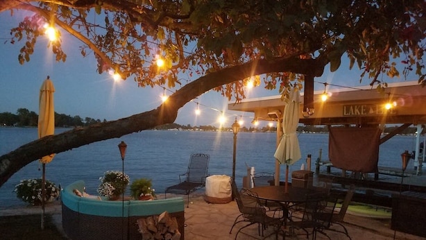 An outdoor seating area with a table and chairs, a covered bench, and a planter with flowers. A large tree provides shade, and a bridge is visible in the background.