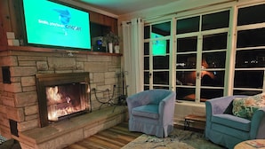 A cozy living room with a stone fireplace, two blue armchairs, a small table, and a large window with a view of the outside.