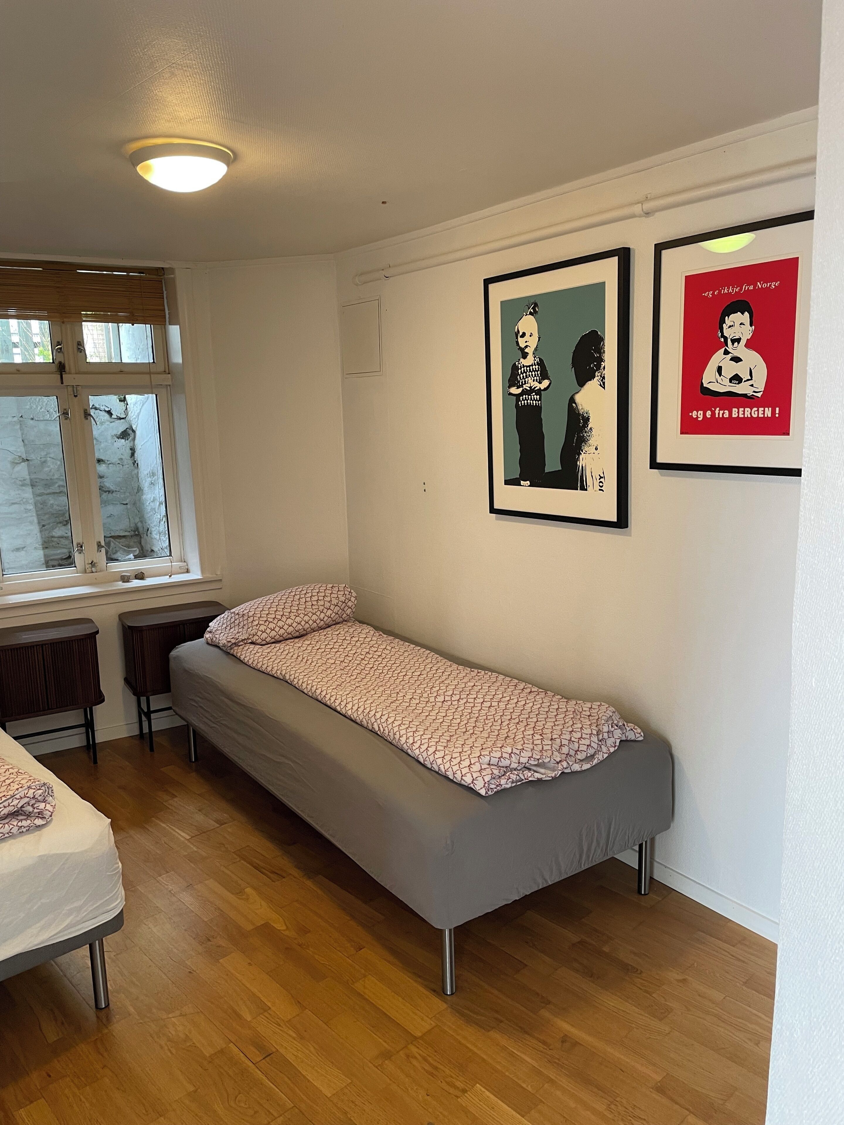 Une chambre avec un lit simple, une fenêtre, un plancher en bois et deux affiches encadrées au mur.