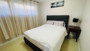 Een net opgemaakt bed met een witte dekbedovertrek en geruite kussens, een donker hoofdbord, een nachtkastje met een lamp en een aan de muur hangend schilderij.
