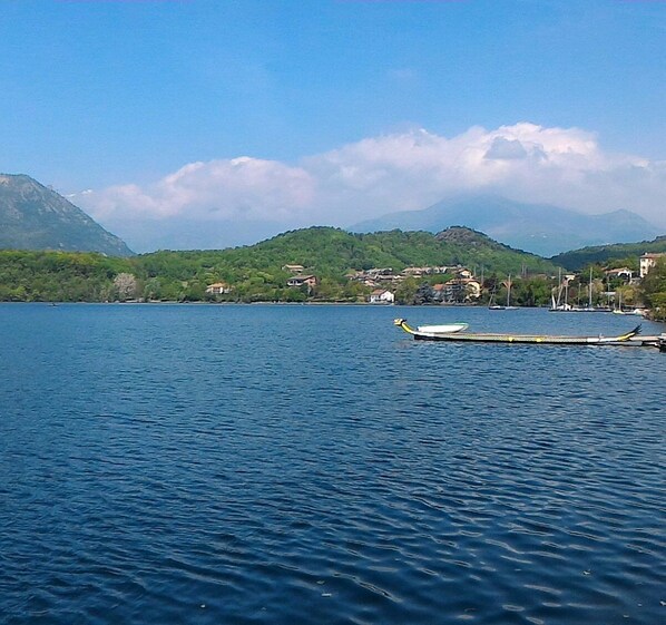 A calm lake with a traditional boat docked on the shore, surrounded by lush green hills and a few buildings in the background.