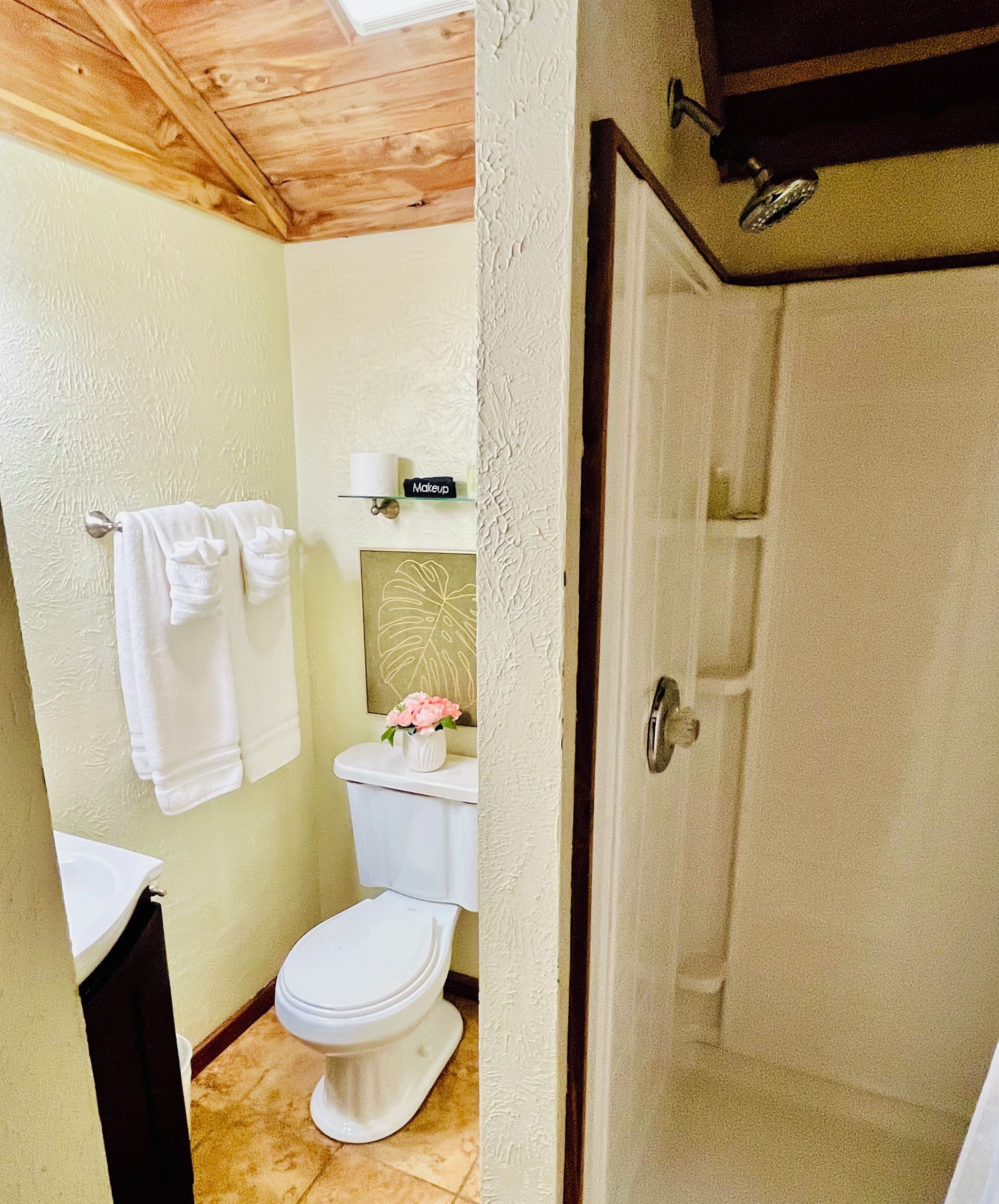 A bathroom with a toilet, sink, and shower. There is a towel rack with towels and a small shelf with a makeup item.