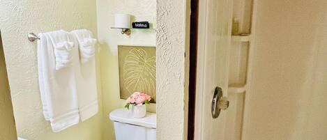A bathroom with a toilet, sink, and shower. There is a towel rack with towels and a small shelf with a makeup item.
