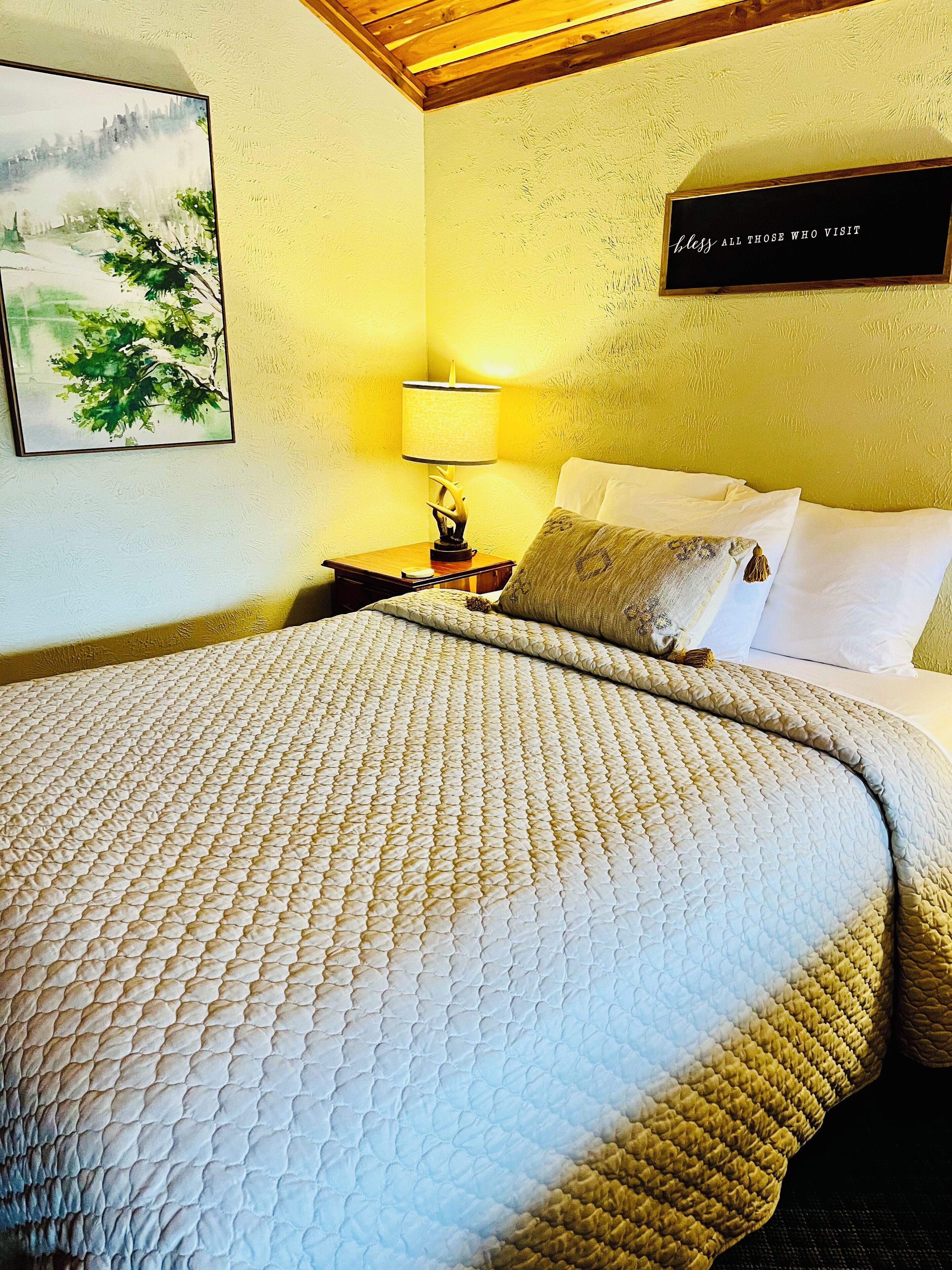 Een net opgemaakt bed met een gewatteerde spreidekbed, een bedlampje, een ingelijste afbeelding en een bordje aan de muur.