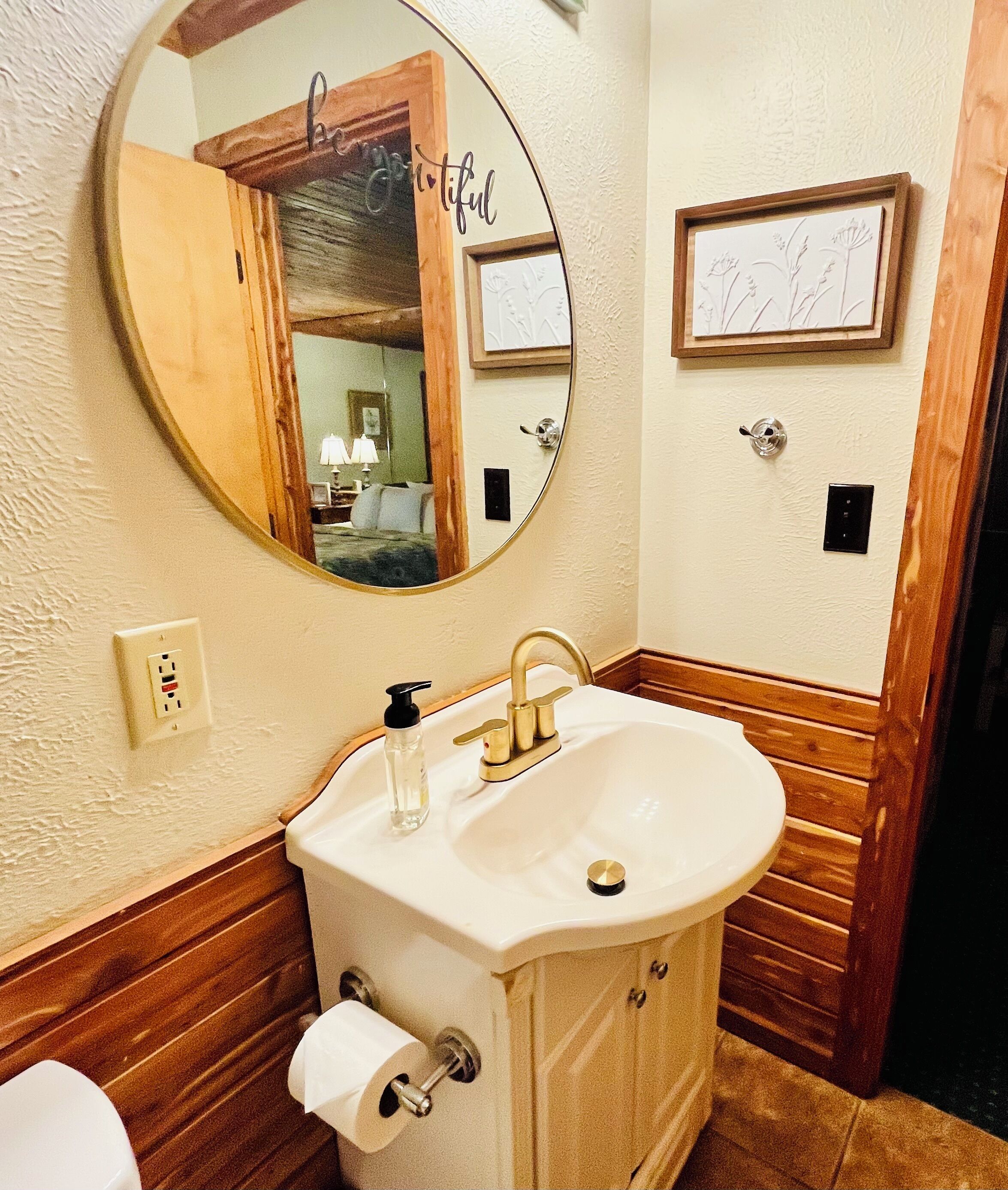 A bathroom with a round mirror, a sink with a gold faucet, wooden paneling, a framed picture on the wall, and a toilet.