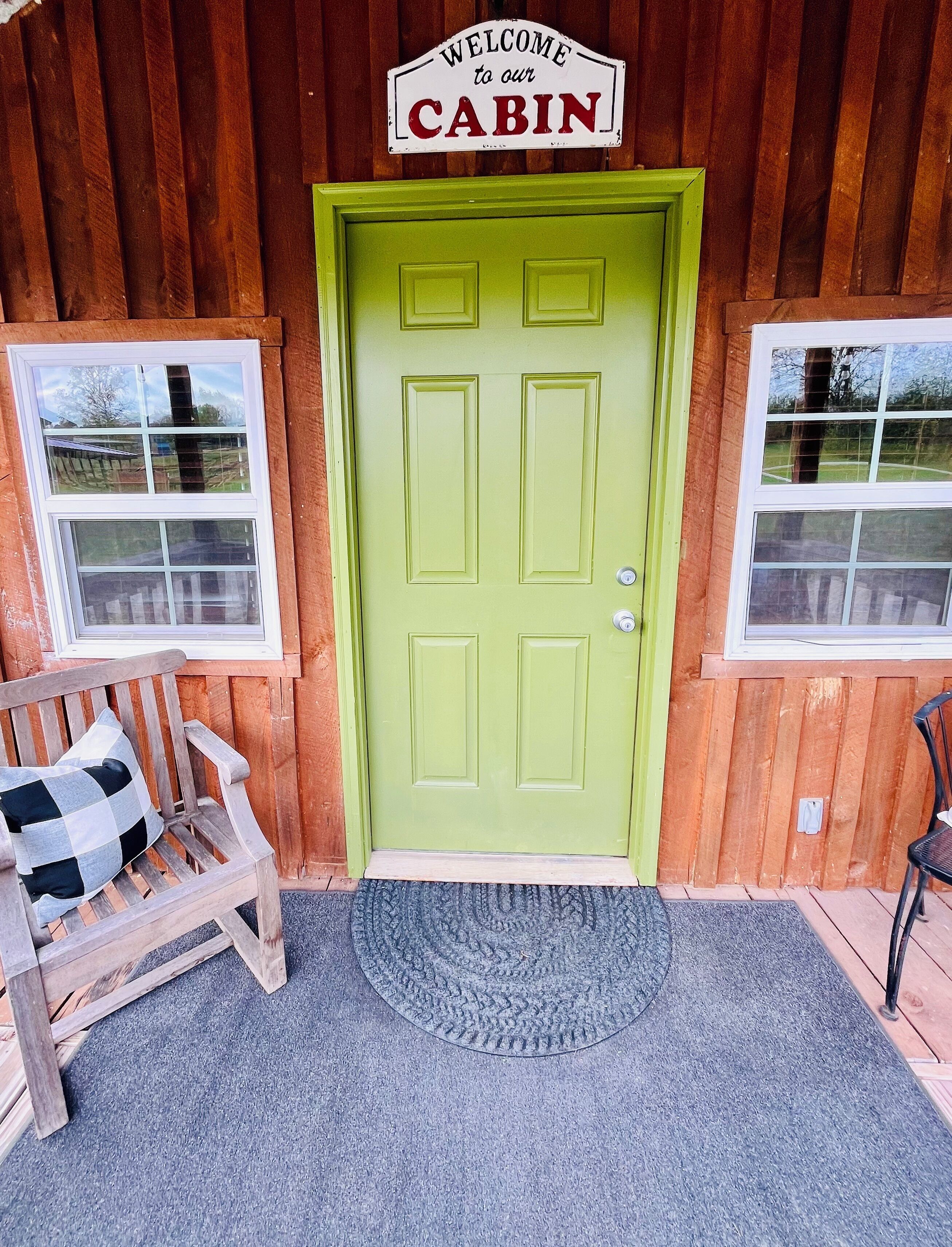 Een groene deur met daar boven een bord waarop staat 'Welkom in onze Houten Hut', een houten bank met een geruit kussen, en een deurmat ervoor.