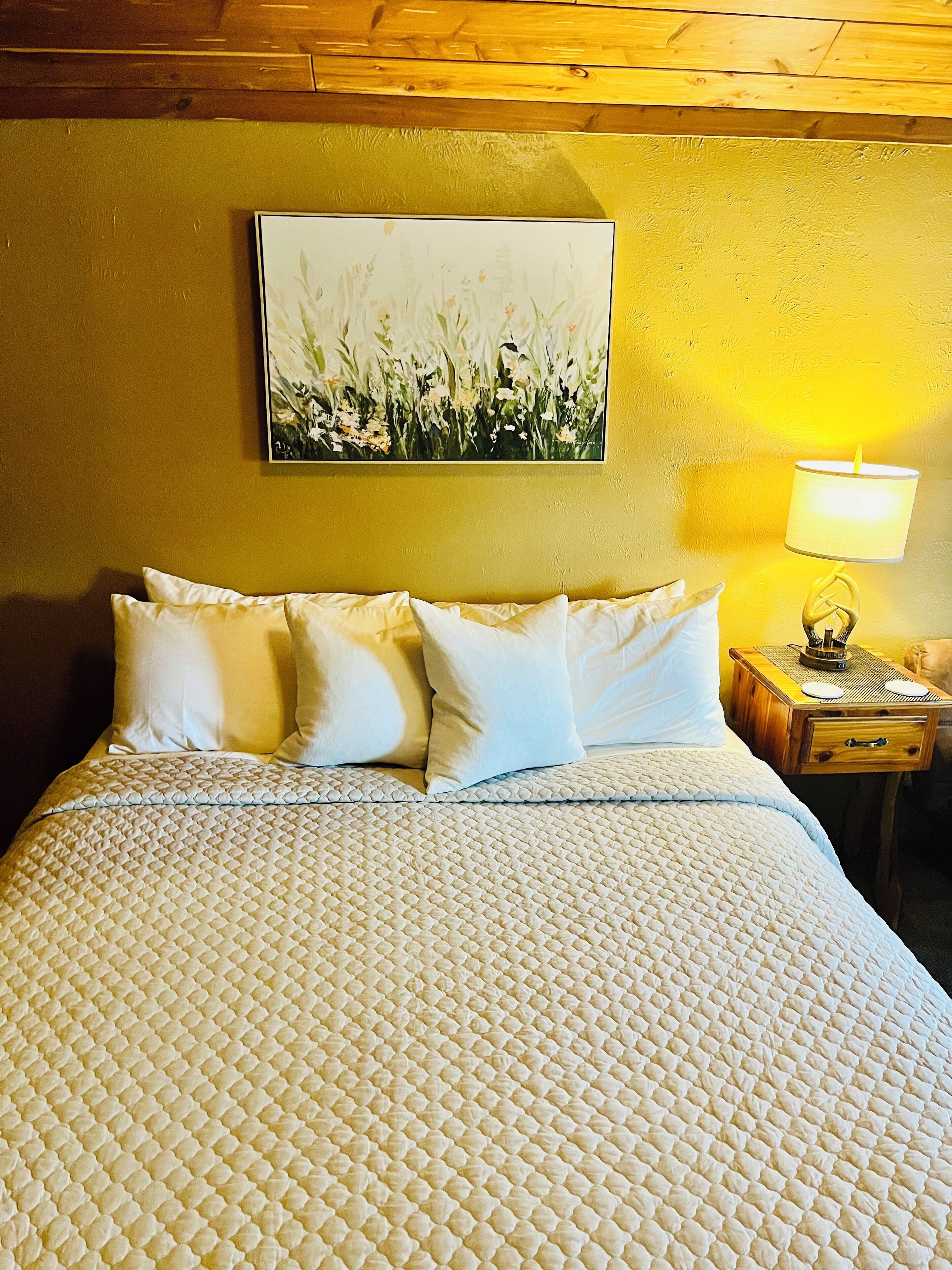 A bed with a quilted cover, a nightstand with a lamp, and a painting on the wall.