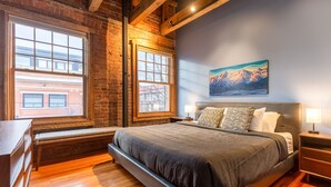 A bedroom with a large bed, a bench, a nightstand, and a painting of mountains on the wall. - Amazing Downtown Location. Sleeps 7. Dog Friendly (Fernie)