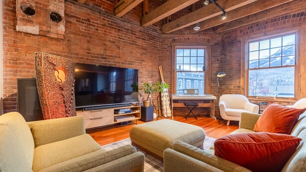 A cozy living room with a brick wall, wooden beams, a flat-screen TV, comfortable seating, and large windows offering a view of the outside. - Amazing Downtown Location. Sleeps 7. Dog Friendly (Fernie)