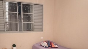 Een compacte, netjes ingerichte slaapkamer met een enkel bed, een nachtkastje, een raam met zonwering en een betegelde vloer.