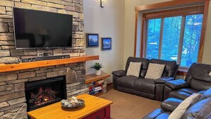 A living room with a stone fireplace, a flat-screen TV mounted above it, a leather sofa, and a window with blinds. - 5 Min. Drive to the Resort & Fernie. Hot Tub (Fernie)