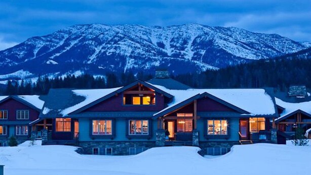 Une rangée de chalets en rondins, aux toits couverts de neige, avec une chaîne de montagnes en arrière-plan.