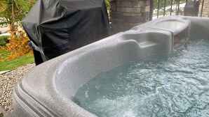 Un bain à remous à l’extérieur, avec un revêtement en pierre, entouré d’une terrasse en pierre et d’un barbecue couvert.
