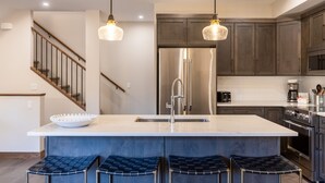 Une cuisine moderne dotée d’un îlot central, d’appareils électroménagers en acier inoxydable, de meubles de cuisine en bois foncé et de luminaires suspendus.