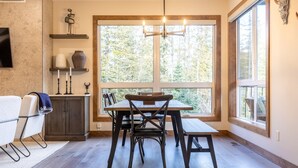 Un coin repas avec une table et des chaises en bois, un lustre et une grande fenêtre donnant sur la forêt.